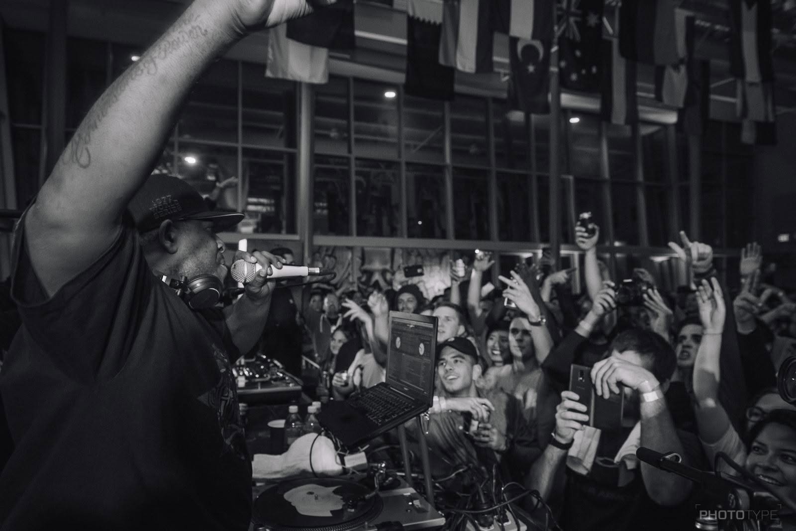 MC performing under a canopy of international flags, arm raised to a cheering crowd