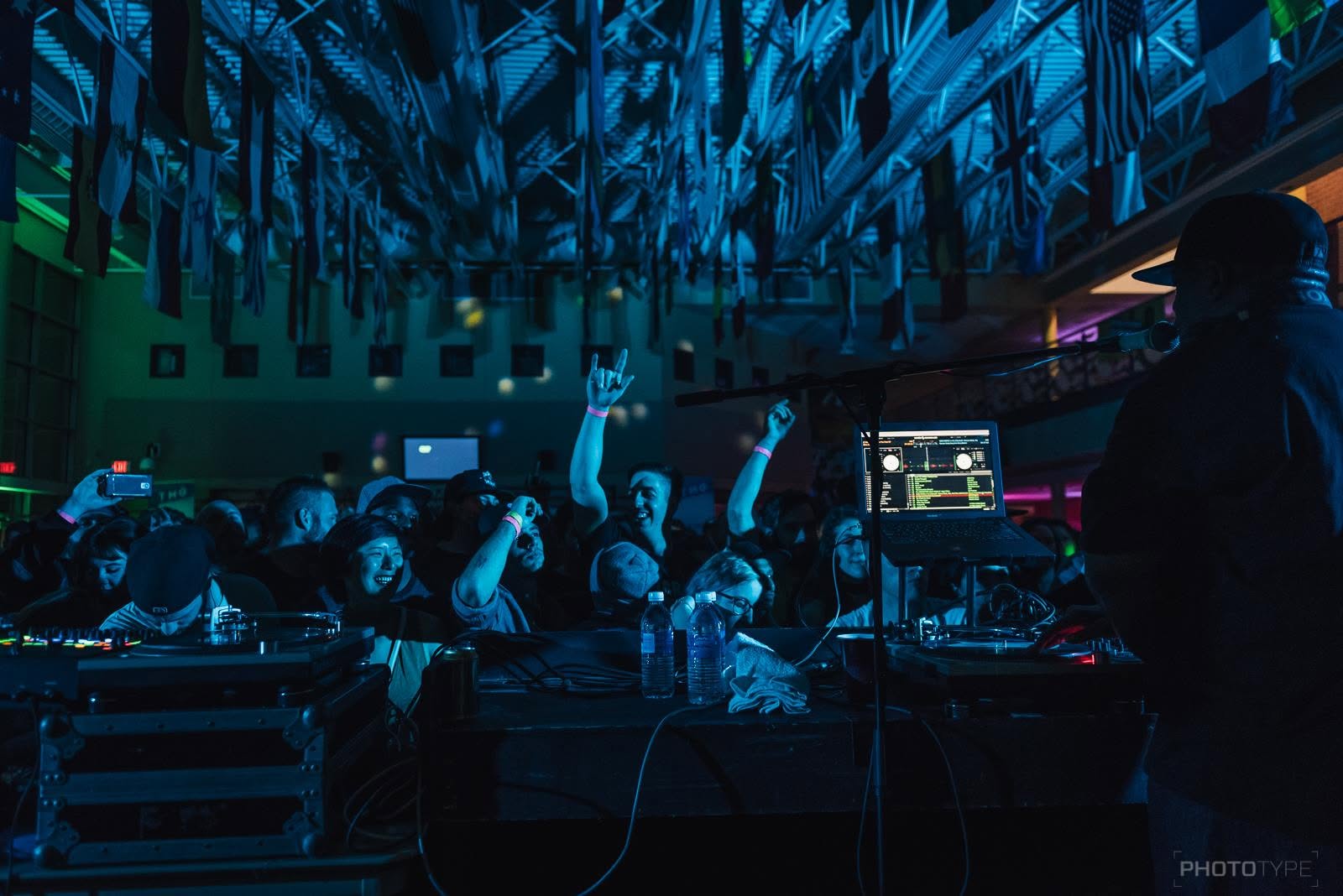 DJ performing to a crowd with hands raised under a flag-draped ceiling