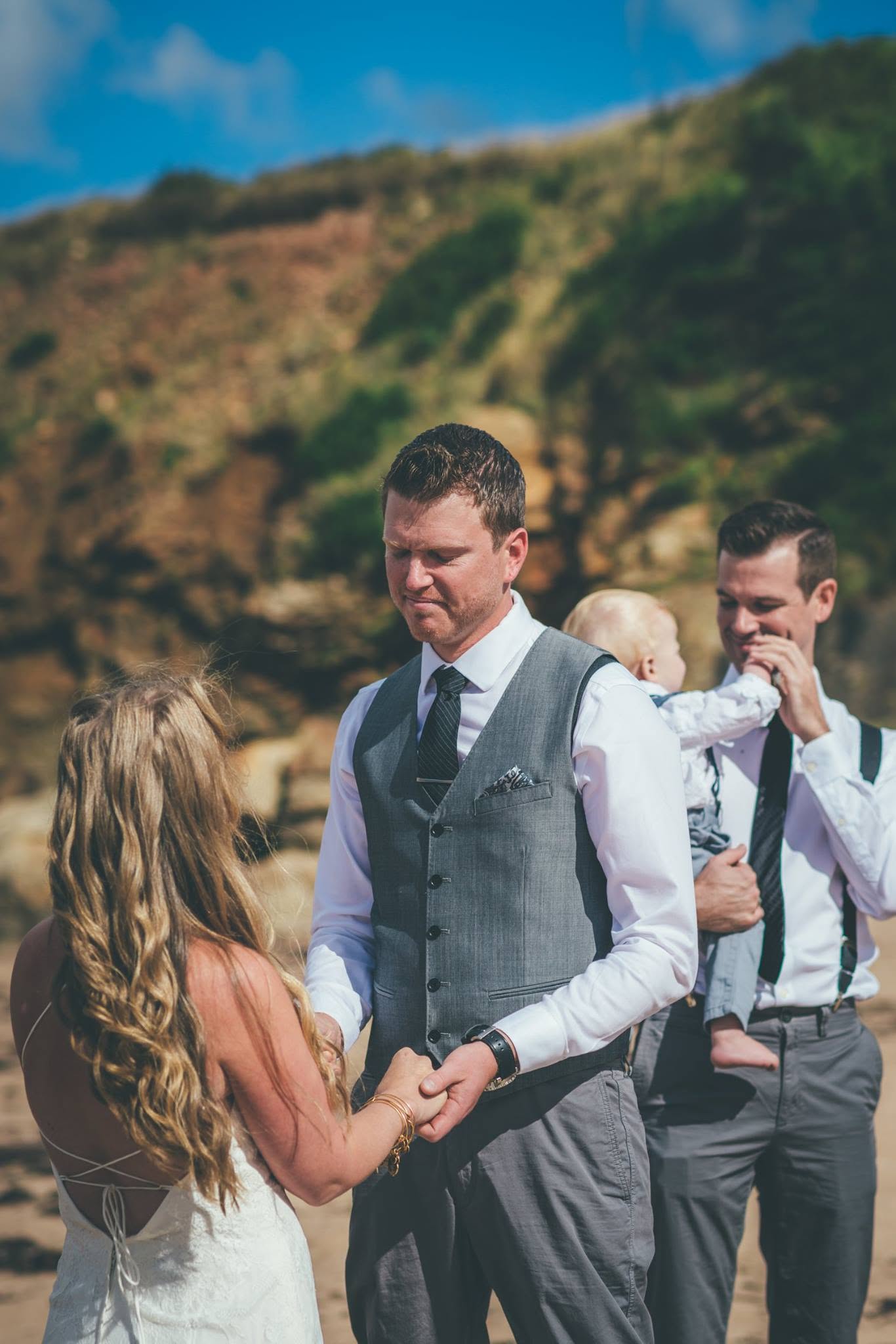 Bride and groom holding hands on the beach, groomsmen and a baby looking on